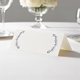 wedding place card with an elegant design on folded white cardstock ready to write your name on. Displayed on a white tablecloth with wine glasses, plates, and a floral centrepiece in the softly blurred background.