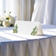 blank wedding place card printed with an elegantdesign on folded white cardstock ready to write your names on. Displayed on a table with a flowing white tablecloth, with soft natural light and white roses in the background.