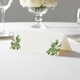 wedding place card with an elegant design on folded white cardstock ready to write your name on. Displayed on a white tablecloth with wine glasses, plates, and a floral centrepiece in the softly blurred background.