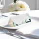 blank wedding place card with elegant design on folded white cardstock. Placed on a white plate with silver cutlery and a neatly folded napkin, with soft-focus white flowers in the background.