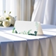 blank wedding place card printed with an elegantdesign on folded white cardstock ready to write your names on. Displayed on a table with a flowing white tablecloth, with soft natural light and white roses in the background.