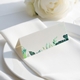 wedding place card with an elegant design on folded white cardstock, ready to write your names on. Positioned on a neatly folded white napkin atop a white plate, with a soft-focus white rose in the background.