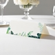 wedding place card with an elegant design on folded white cardstock ready to write your name on. Displayed on a white tablecloth with wine glasses, plates, and a floral centrepiece in the softly blurred background.