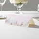 wedding place card with an elegant design on folded white cardstock ready to write your name on. Displayed on a white tablecloth with wine glasses, plates, and a floral centrepiece in the softly blurred background.