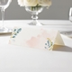 wedding place card with an elegant design on folded white cardstock ready to write your name on. Displayed on a white tablecloth with wine glasses, plates, and a floral centrepiece in the softly blurred background.