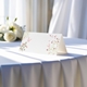 blank wedding place card printed with an elegantdesign on folded white cardstock ready to write your names on. Displayed on a table with a flowing white tablecloth, with soft natural light and white roses in the background.