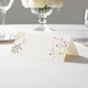 wedding place card with an elegant design on folded white cardstock ready to write your name on. Displayed on a white tablecloth with wine glasses, plates, and a floral centrepiece in the softly blurred background.