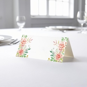 blank wedding place card with an elegant design printed on folded white cardstock, ready to write your names. Placed on a table set for a formal event, with plates, cutlery, and wine glasses in the softly blurred background.