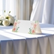 blank wedding place card printed with an elegant design on folded white cardstock ready to write your names on. Displayed on a table with a flowing white tablecloth, with soft natural light and white roses in the background.