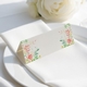 wedding place card with an elegant design on folded white cardstock, ready to write your names on. Positioned on a neatly folded white napkin atop a white plate, with a soft-focus white rose in the background.