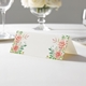 wedding place card with an elegant design on folded white cardstock ready to write your name on. Displayed on a white tablecloth with wine glasses, plates, and a floral centrepiece in the softly blurred background.