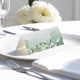 blank wedding place card with elegant design on folded white cardstock. Placed on a white plate with silver cutlery and a neatly folded napkin, with soft-focus white flowers in the background.