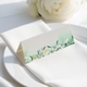 wedding place card with an elegant design on folded white cardstock, ready to write your names on. Positioned on a neatly folded white napkin atop a white plate, with a soft-focus white rose in the background.