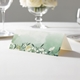 wedding place card with an elegant design on folded white cardstock ready to write your name on. Displayed on a white tablecloth with wine glasses, plates, and a floral centrepiece in the softly blurred background.