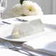 blank wedding place card with elegant design on folded white cardstock. Placed on a white plate with silver cutlery and a neatly folded napkin, with soft-focus white flowers in the background.