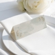 wedding place card with an elegant design on folded white cardstock, ready to write your names on. Positioned on a neatly folded white napkin atop a white plate, with a soft-focus white rose in the background.