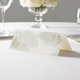 wedding place card with an elegant design on folded white cardstock ready to write your name on. Displayed on a white tablecloth with wine glasses, plates, and a floral centrepiece in the softly blurred background.