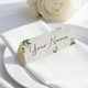 Personalised wedding place card with your name in elegant design on folded white cardstock. Positioned on a neatly folded white napkin atop a white plate, with a soft-focus white rose in the background.