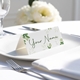 Personalised wedding place card with your name in elegant designt on folded white cardstock. Placed on a white plate with silver cutlery and a neatly folded napkin, with soft-focus white flowers in the background.