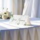 Personalised wedding place card with your name in elegant design on folded white cardstock. Displayed on a table with a flowing white tablecloth, with soft natural light and white roses in the background.