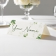 Personalised wedding place card with your name in elegant design on folded white cardstock. Displayed on a white tablecloth with wine glasses, plates, and a floral centrepiece in the softly blurred background.