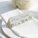 Personalised wedding place card with your name in elegant design on folded white cardstock. Positioned on a neatly folded white napkin atop a white plate, with a soft-focus white rose in the background.