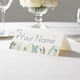 Personalised wedding place card with your name in elegant design on folded white cardstock. Displayed on a white tablecloth with wine glasses, plates, and a floral centrepiece in the softly blurred background.