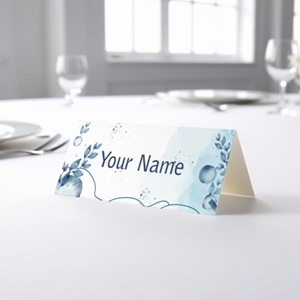 Personalised wedding place card with your name in elegant design on folded white cardstock. Placed on a table set for a formal event, with plates, cutlery, and wine glasses in the softly blurred background.