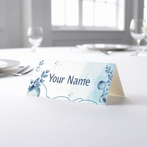 Personalised wedding place card with your name in elegant design on folded white cardstock. Placed on a table set for a formal event, with plates, cutlery, and wine glasses in the softly blurred background.