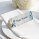 Personalised wedding place card with your name in elegant design on folded white cardstock. Positioned on a neatly folded white napkin atop a white plate, with a soft-focus white rose in the background.