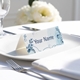 Personalised wedding place card with your name in elegant designt on folded white cardstock. Placed on a white plate with silver cutlery and a neatly folded napkin, with soft-focus white flowers in the background.