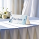 Personalised wedding place card with your name in elegant design on folded white cardstock. Displayed on a table with a flowing white tablecloth, with soft natural light and white roses in the background.