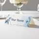 Personalised wedding place card with your name in elegant design on folded white cardstock. Displayed on a white tablecloth with wine glasses, plates, and a floral centrepiece in the softly blurred background.