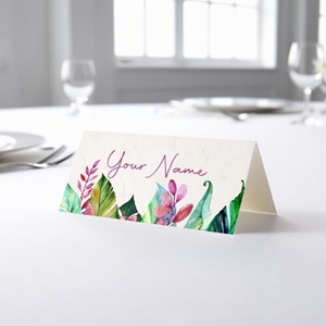 Personalised wedding place card with your name in elegant design on folded white cardstock. Placed on a table set for a formal event, with plates, cutlery, and wine glasses in the softly blurred background.