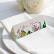 Personalised wedding place card with your name in elegant design on folded white cardstock. Positioned on a neatly folded white napkin atop a white plate, with a soft-focus white rose in the background.