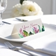 Personalised wedding place card with your name in elegant designt on folded white cardstock. Placed on a white plate with silver cutlery and a neatly folded napkin, with soft-focus white flowers in the background.