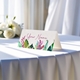 Personalised wedding place card with your name in elegant design on folded white cardstock. Displayed on a table with a flowing white tablecloth, with soft natural light and white roses in the background.