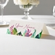 Personalised wedding place card with your name in elegant design on folded white cardstock. Displayed on a white tablecloth with wine glasses, plates, and a floral centrepiece in the softly blurred background.