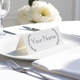 Personalised wedding place card with your name in elegant designt on folded white cardstock. Placed on a white plate with silver cutlery and a neatly folded napkin, with soft-focus white flowers in the background.