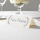 Personalised wedding place card with your name in elegant design on folded white cardstock. Displayed on a white tablecloth with wine glasses, plates, and a floral centrepiece in the softly blurred background.