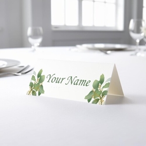 Personalised wedding place card with your name in elegant design on folded white cardstock. Placed on a table set for a formal event, with plates, cutlery, and wine glasses in the softly blurred background.