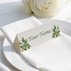 Personalised wedding place card with your name in elegant design on folded white cardstock. Positioned on a neatly folded white napkin atop a white plate, with a soft-focus white rose in the background.