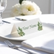 Personalised wedding place card with your name in elegant designt on folded white cardstock. Placed on a white plate with silver cutlery and a neatly folded napkin, with soft-focus white flowers in the background.