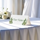 Personalised wedding place card with your name in elegant design on folded white cardstock. Displayed on a table with a flowing white tablecloth, with soft natural light and white roses in the background.