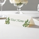 Personalised wedding place card with your name in elegant design on folded white cardstock. Displayed on a white tablecloth with wine glasses, plates, and a floral centrepiece in the softly blurred background.