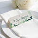 Personalised wedding place card with your name in elegant design on folded white cardstock. Positioned on a neatly folded white napkin atop a white plate, with a soft-focus white rose in the background.