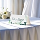 Personalised wedding place card with your name in elegant design on folded white cardstock. Displayed on a table with a flowing white tablecloth, with soft natural light and white roses in the background.