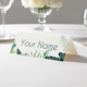 Personalised wedding place card with your name in elegant design on folded white cardstock. Displayed on a white tablecloth with wine glasses, plates, and a floral centrepiece in the softly blurred background.