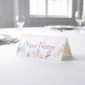 Personalised wedding place card with your name in elegant design on folded white cardstock. Placed on a table set for a formal event, with plates, cutlery, and wine glasses in the softly blurred background.