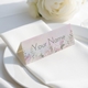 Personalised wedding place card with your name in elegant design on folded white cardstock. Positioned on a neatly folded white napkin atop a white plate, with a soft-focus white rose in the background.